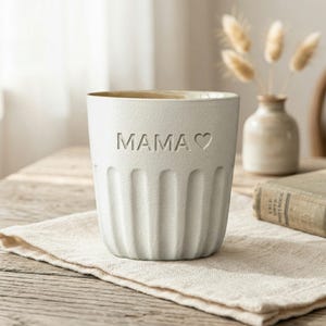 Puede incluir: Una taza de cerámica blanca con la palabra "MAMA" y un símbolo de corazón en relieve en la parte delantera. La taza tiene ranuras verticales y está sobre una tela beige. Un pequeño jarrón con flores secas está en el fondo.