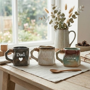 Könnte beinhalten: Drei Keramikbecher mit einzigartigen Designs stehen auf einem Holztisch. Ein Becher ist dunkelgrau mit "Dad" und einem Herz, ein anderer ist beige mit "Mamaa" und einem Herz, und der dritte ist mehrfarbig mit "Olivia" und "EST. 2026".