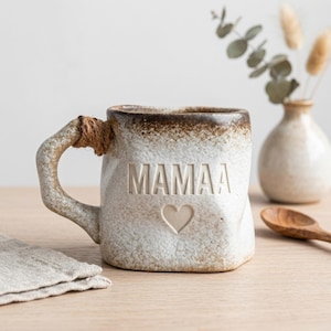 May include: A ceramic mug with the word "MAMAA" and a heart etched on the front. The mug has a textured, off-white finish with a brown rim and handle. The handle is wrapped with twine. The mug is on a wooden surface.