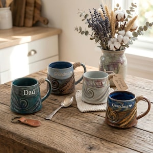 May include: Four ceramic mugs with rope handles and the words "Dad", "Mom", and "Grandma" in white lettering. The mugs are various colours, including teal, blue, and brown. A spoon and leather keyring are on the wooden table.