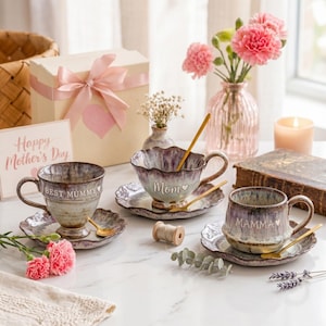 May include: A Mother's Day still life featuring three decorative teacups and saucers with gold spoons. The teacups are inscribed with "Best Mummy", "Mom", and "Mamma". Pink carnations and a gift box with a pink bow are also present.