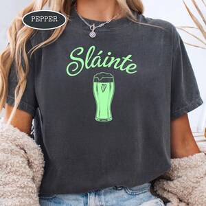 K&ouml;nnte beinhalten: Dunkelgraues T-Shirt mit dem Wort "Sl&aacute;inte" in gr&uuml;ner Schrift &uuml;ber einer gr&uuml;nen Bierglas-Grafik. Das Shirt hat eine lockere Passform und ist aus einem weichen Material. Das Wort "Pepper" befindet sich in einem wei&szlig;en Oval in der oberen linken Ecke.