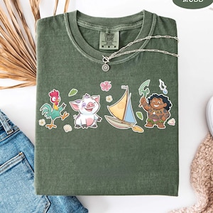 May include: Moss green t-shirt with a crew neck and a graphic of cartoon characters. The graphic includes a rooster, a pig, a sailboat, and a character holding a weapon. A silver necklace with a pendant is draped on the shirt.