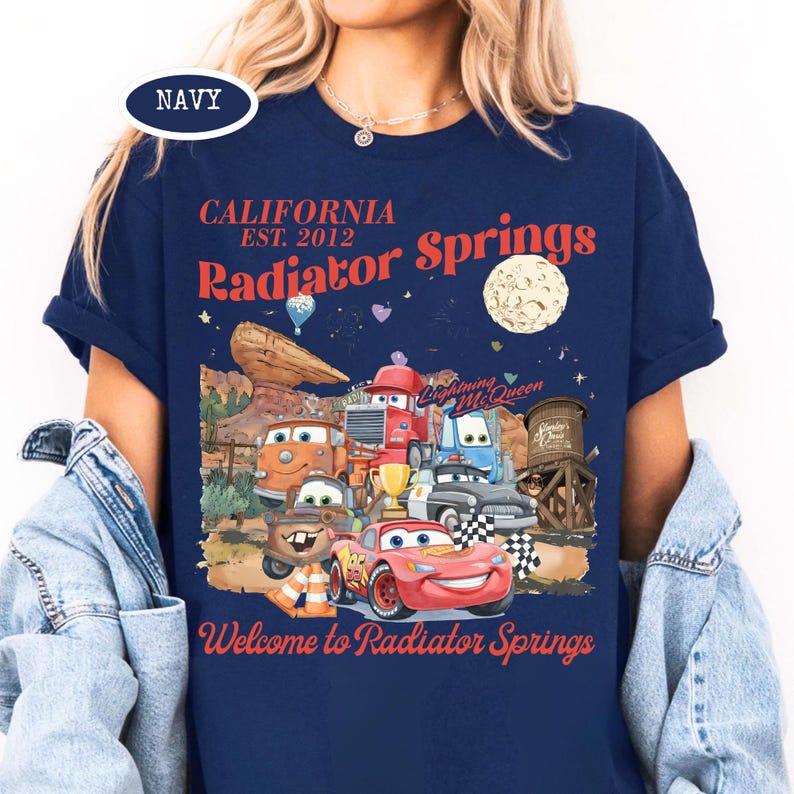 May include: Navy blue t-shirt featuring a graphic of characters from the movie Cars, with the text "CALIFORNIA EST. 2012 Radiator Springs" and "Welcome to Radiator Springs". The design includes a moon, desert landscape, and various car characters.