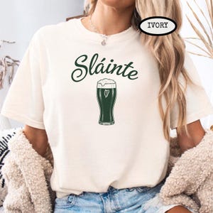 K&ouml;nnte beinhalten: Elfenbeinfarbenes T-Shirt mit dem Wort "Sl&aacute;inte" in gr&uuml;ner Schrift &uuml;ber einem gr&uuml;nen Bierkrug mit herzf&ouml;rmigem Schaumdesign. Das Shirt ist ein l&auml;ssiger Stil.
