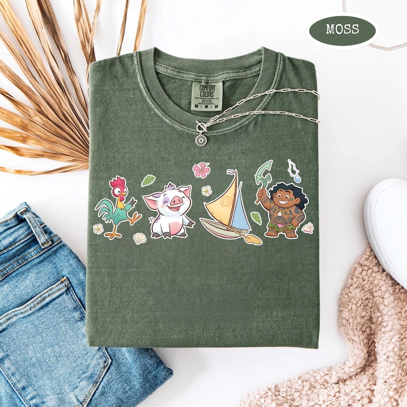 May include: Moss green t-shirt with a crew neck and a graphic of cartoon characters. The graphic includes a rooster, a pig, a sailboat, and a character holding a weapon. A silver necklace with a pendant is draped on the shirt.