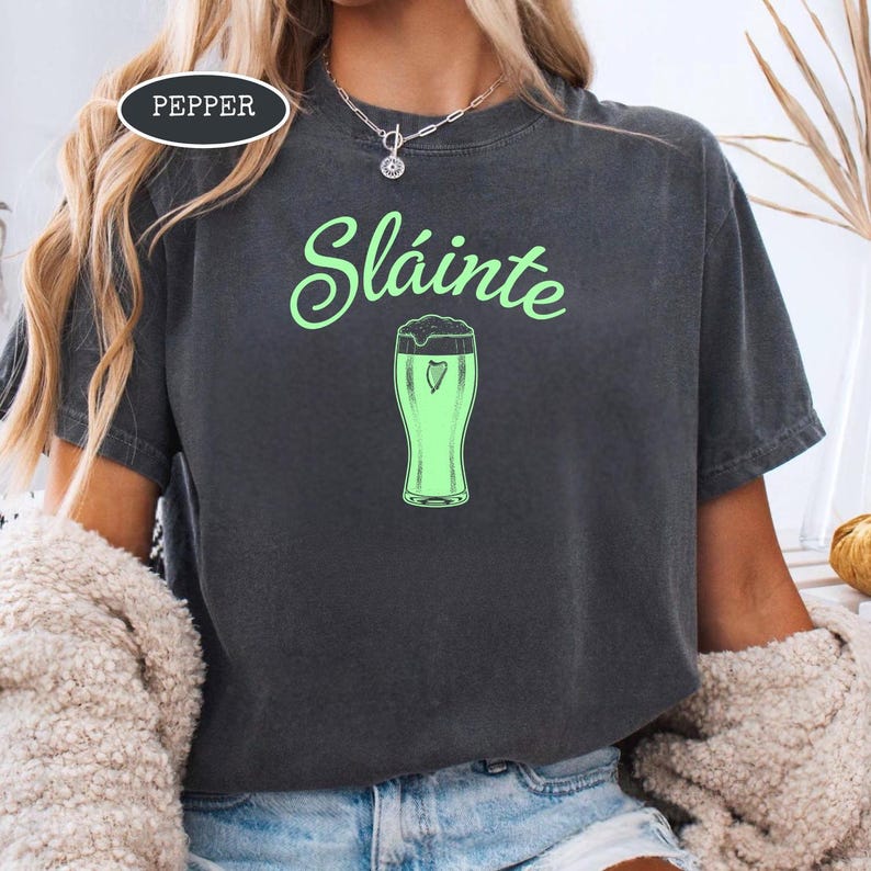 K&ouml;nnte beinhalten: Dunkelgraues T-Shirt mit dem Wort "Sl&aacute;inte" in gr&uuml;ner Schrift &uuml;ber einer gr&uuml;nen Bierglas-Grafik. Das Shirt hat eine lockere Passform und ist aus einem weichen Material. Das Wort "Pepper" befindet sich in einem wei&szlig;en Oval in der oberen linken Ecke.