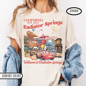 May include: Ivory-colored t-shirt featuring a graphic of characters from the movie Cars, including Lightning McQueen. The design includes the text "CALIFORNIA EST. 2012 Radiator Springs" and "Welcome to Radiator Springs."