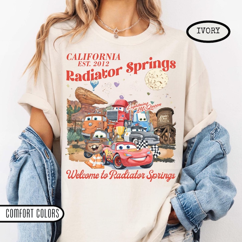 May include: Ivory-colored t-shirt featuring a graphic of characters from the movie Cars, including Lightning McQueen. The design includes the text "CALIFORNIA EST. 2012 Radiator Springs" and "Welcome to Radiator Springs."