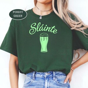 K&ouml;nnte beinhalten: Forstgr&uuml;nes T-Shirt mit dem Wort "Sl&aacute;inte" in hellgr&uuml;ner Schrift &uuml;ber einer hellgr&uuml;nen Bierglas-Illustration. Das Bierglas hat eine Herzform im Schaum. Das Shirt hat eine einfarbige forstgr&uuml;ne Farbe.