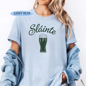 K&ouml;nnte beinhalten: Hellblaues T-Shirt mit dem Wort "Sl&aacute;inte" in dunkelgr&uuml;ner Schrift &uuml;ber einem gr&uuml;nen Bierkrug mit wei&szlig;em Schaum. Das Shirt hat kurze &Auml;rmel und einen Rundhalsausschnitt. Der Text "LIGHT BLUE" befindet sich in einem hellblauen Feld.