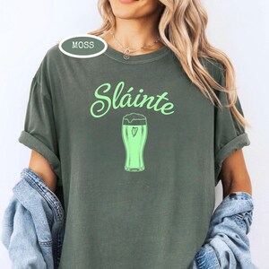 K&ouml;nnte beinhalten: Moosgr&uuml;nes T-Shirt mit dem Wort "Sl&aacute;inte" in hellgr&uuml;ner Schrift &uuml;ber einem hellgr&uuml;nen Bierkrug mit einem Herz. Das Wort "Moss" befindet sich in einem gr&uuml;nen Oval oben links.