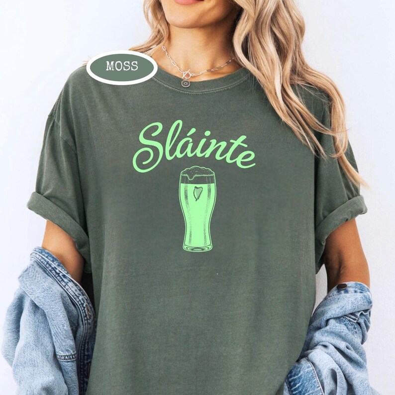 K&ouml;nnte beinhalten: Moosgr&uuml;nes T-Shirt mit dem Wort "Sl&aacute;inte" in hellgr&uuml;ner Schrift &uuml;ber einem hellgr&uuml;nen Bierkrug mit einem Herz. Das Wort "Moss" befindet sich in einem gr&uuml;nen Oval oben links.