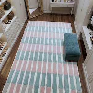 May include: A patterned rug featuring vertical, curved shapes in pink, teal, and grey. The rug is laid on a dark wooden floor. A blue upholstered bench sits nearby. The room is well-lit and has a large mirror.