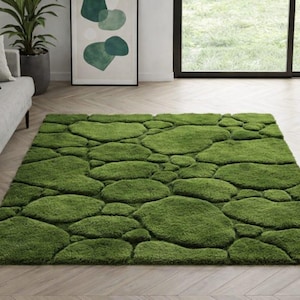 May include: A rectangular green rug with a pebble-like design. The rug is made of a soft, plush material and is placed on a light-colored wooden floor. A gray sofa and green pillows are in the background.
