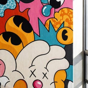 May include: A colorful abstract rug with a cartoonish design. The rug features a white face with crossed-out eyes and a pink nose, surrounded by orange, pink, blue, and black shapes. The rug is rectangular and appears to be made of a soft material.