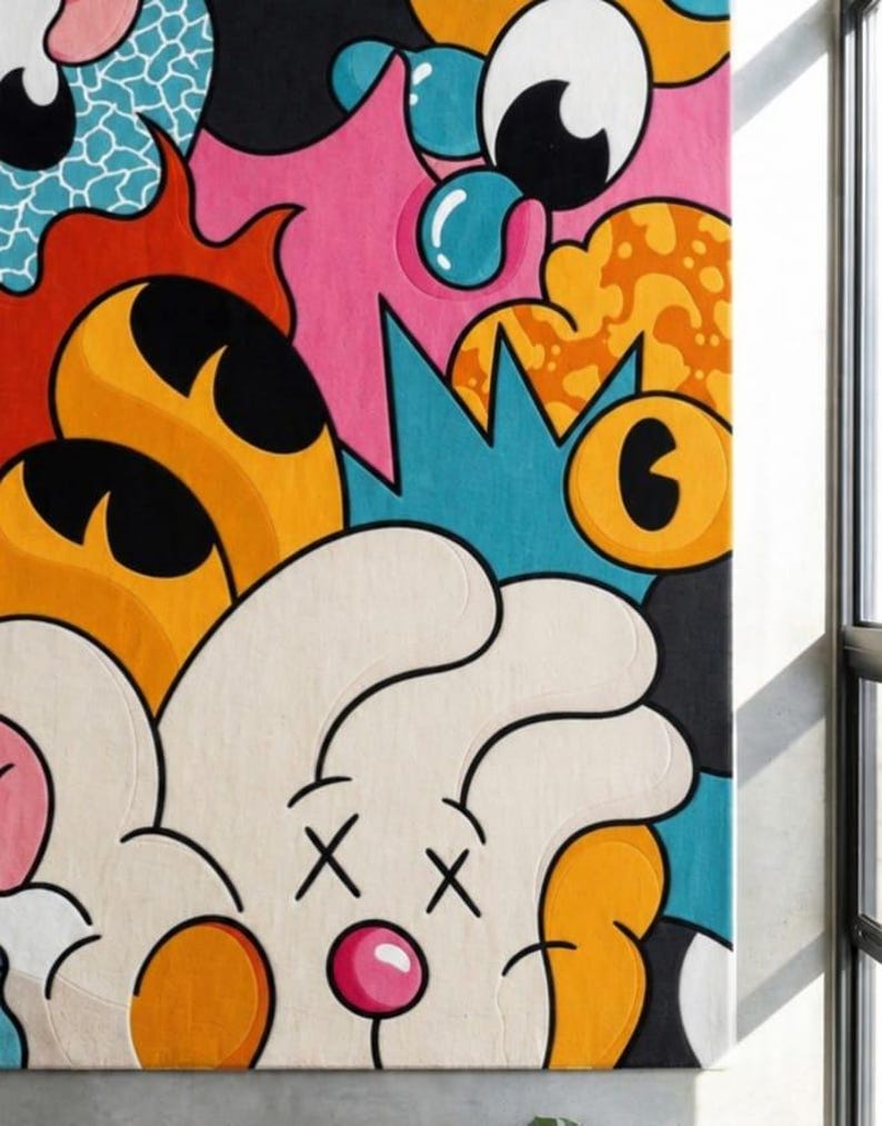 May include: A colorful abstract rug with a cartoonish design. The rug features a white face with crossed-out eyes and a pink nose, surrounded by orange, pink, blue, and black shapes. The rug is rectangular and appears to be made of a soft material.