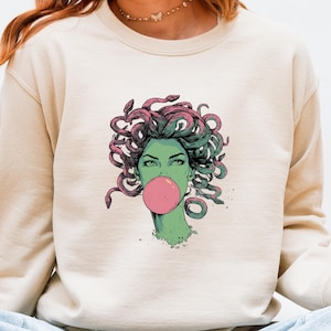 May include: Beige sweatshirt featuring a graphic of Medusa blowing a pink bubble. The Medusa illustration has green skin, pink and green snakes for hair, and is set against a neutral background. The sweatshirt is a crew neck style.