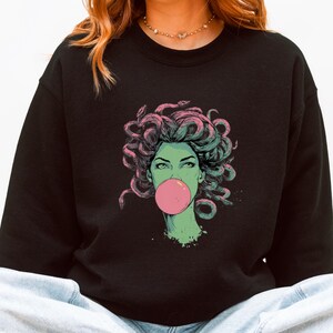 May include: Black sweatshirt featuring a graphic of Medusa with snakes for hair, blowing a pink bubble. The Medusa illustration has green skin and is outlined in pink and green. The sweatshirt is a crew neck.