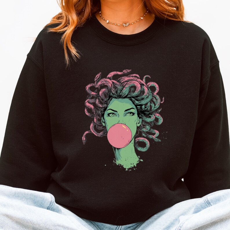 May include: Black sweatshirt featuring a graphic of Medusa with snakes for hair, blowing a pink bubble. The Medusa illustration has green skin and is outlined in pink and green. The sweatshirt is a crew neck.