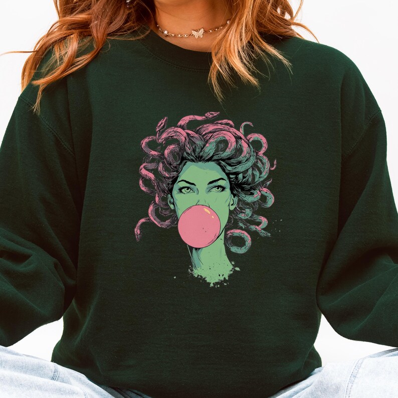 May include: Dark green sweatshirt featuring a graphic of a woman with snake hair blowing a pink bubble. The woman's skin is green, and her hair is pink and gray. The sweatshirt is a solid dark green color.