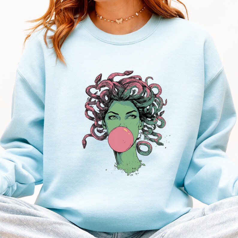 May include: Light blue sweatshirt featuring a graphic of Medusa blowing a bubblegum bubble. The Medusa illustration has green skin, green eyes, and pink and green snakes for hair. The bubble is pink.