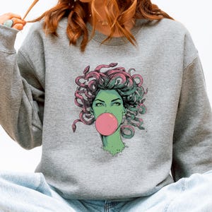 May include: A gray sweatshirt featuring a graphic of Medusa with green skin, pink bubblegum, and snakes for hair. The Medusa illustration has green eyes and is set against a gray background. The sweatshirt is a casual style.