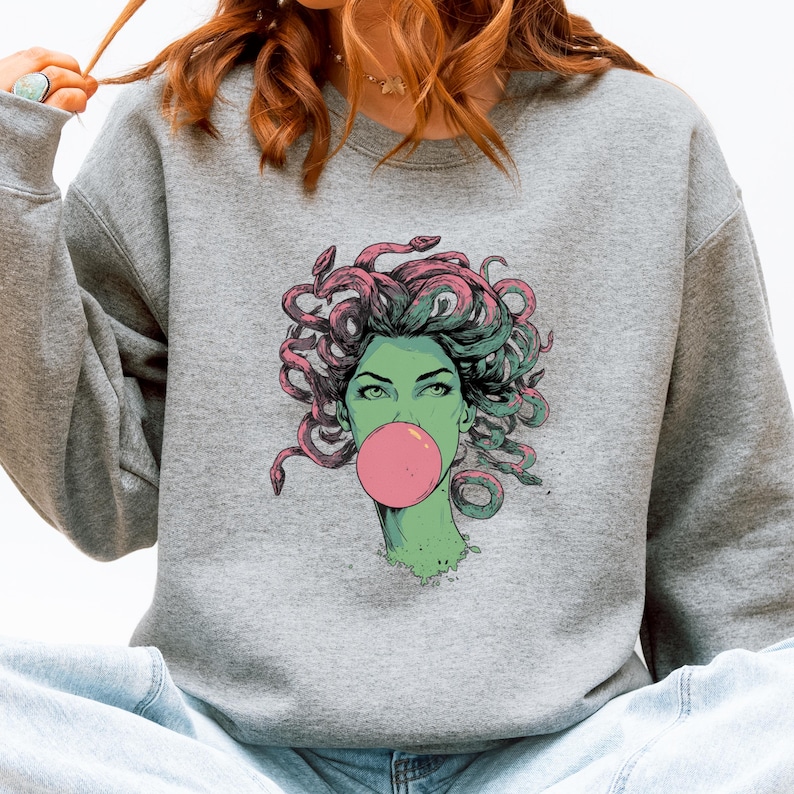 May include: A gray sweatshirt featuring a graphic of Medusa with green skin, pink bubblegum, and snakes for hair. The Medusa illustration has green eyes and is set against a gray background. The sweatshirt is a casual style.