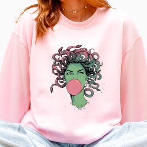May include: A light pink sweatshirt featuring a graphic of Medusa with green skin, snakes for hair, and a pink bubble gum bubble. The illustration is detailed with a pop art style. The sweatshirt is a solid color.
