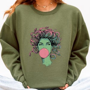 May include: Olive green sweatshirt featuring a graphic of a woman with snakes for hair blowing a pink bubble. The illustration has a teal and pink color scheme. The sweatshirt is a crew neck style.