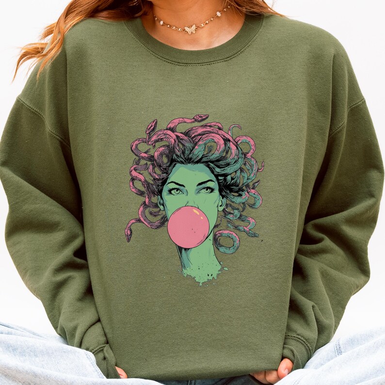 May include: Olive green sweatshirt featuring a graphic of a woman with snakes for hair blowing a pink bubble. The illustration has a teal and pink color scheme. The sweatshirt is a crew neck style.