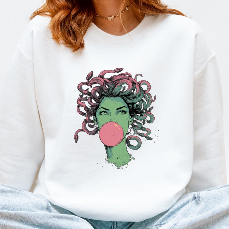 May include: White sweatshirt featuring a graphic of Medusa with green skin, pink hair, and a pink bubblegum bubble. The illustration has a comic book style. The sweatshirt is a casual, comfortable garment.