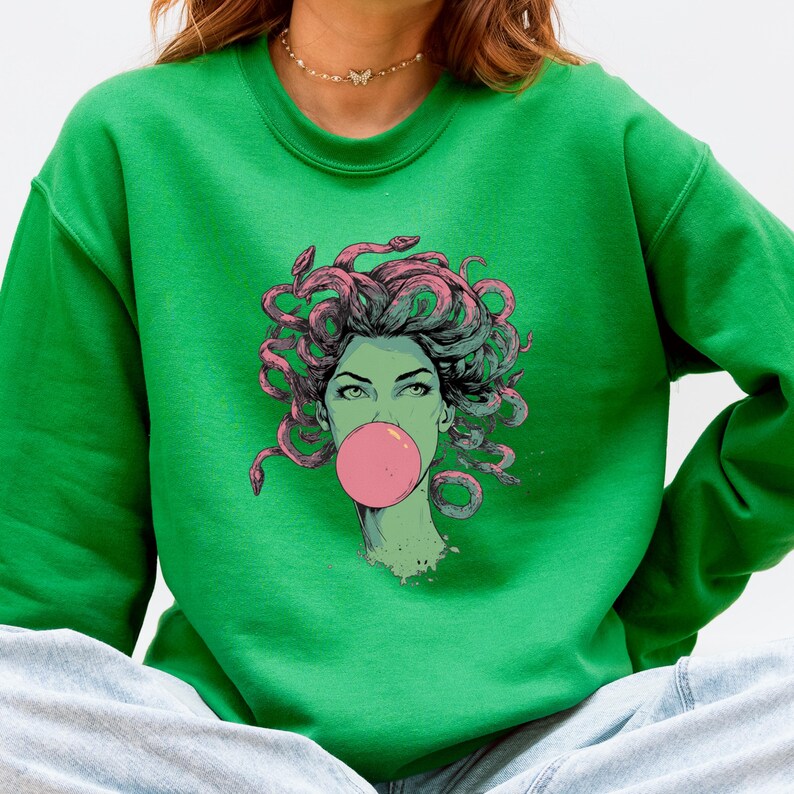 May include: Green sweatshirt featuring a graphic of Medusa blowing a bubblegum bubble. The Medusa illustration has green skin, pink hair, and snakes. The sweatshirt is a solid green color.
