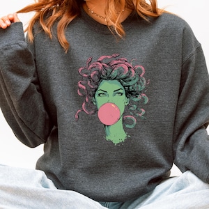 May include: Dark gray sweatshirt featuring a graphic of a woman with green skin, pink hair of snakes, and a pink bubble gum bubble. The illustration has a comic book style. The sweatshirt is a crew neck.