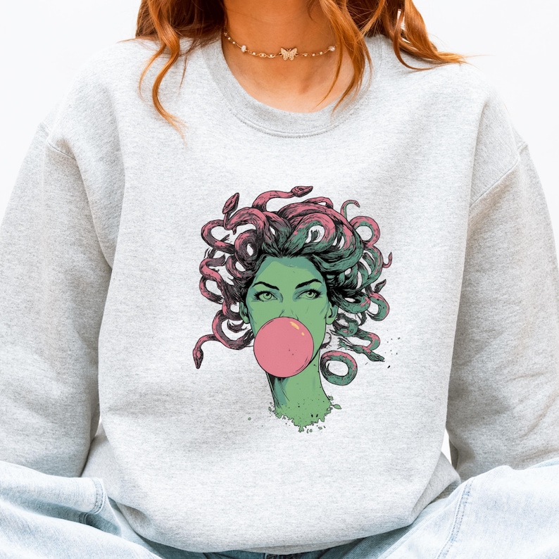 May include: A light gray sweatshirt featuring a graphic of Medusa with green skin, pink bubblegum, and snakes for hair. The Medusa illustration is in a comic book style. The sweatshirt is a casual, comfortable style.
