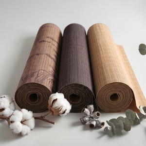 May include: Three cylindrical rolls of textured material arranged side by side. The left roll is medium brown with a dark line pattern, the center roll is dark brown, and the right roll is light tan. White cotton bolls and eucalyptus pods sit nearby.