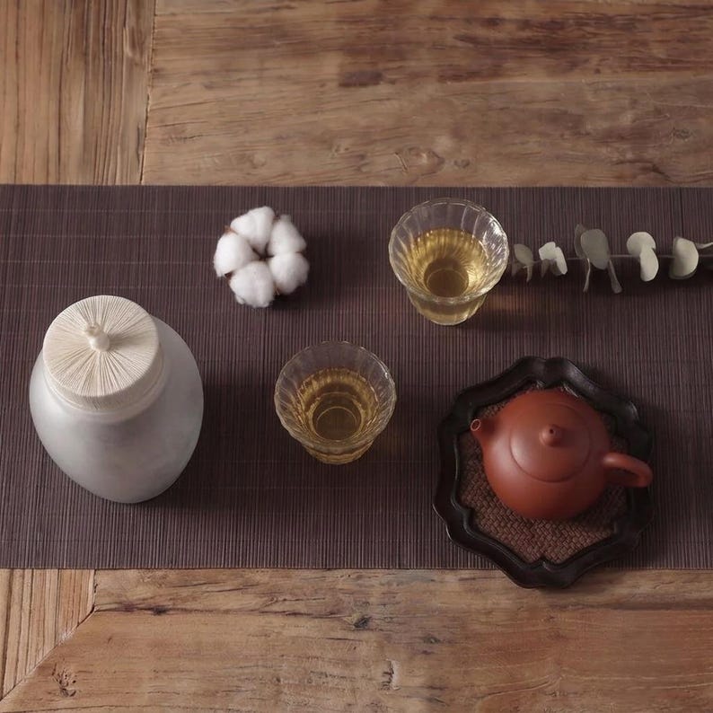 May include: A white ceramic tea jar with a ribbed lid, two clear glass cups filled with light yellow tea, a small brown clay teapot on a dark scalloped tray, a white cotton flower, and a sprig of dried eucalyptus leaves on a dark mat.