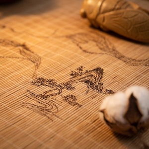 May include: A light brown bamboo mat featuring a dark brown pyrography illustration of a landscape with trees and mountains. A carved wooden cicada figurine and a dried cotton boll are placed on the textured surface of the bamboo matting.