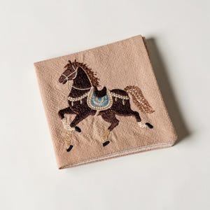 May include: A folded, tan-colored textile featuring an embroidered horse design. The horse is brown with a blue saddle and tan and gold accents. The embroidery is detailed, and the textile has a textured appearance.