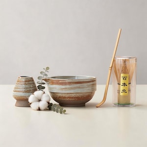 May include: A ceramic matcha bowl, whisk, and accessories set. The bowl is brown and white striped. A bamboo whisk is in a clear container with Japanese text. A bamboo scoop and a small ceramic vase are also included.