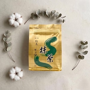 May include: A gold foil package of matcha tea with Japanese characters and a green brushstroke design. The package is surrounded by cotton buds and eucalyptus leaves on a light-colored surface.