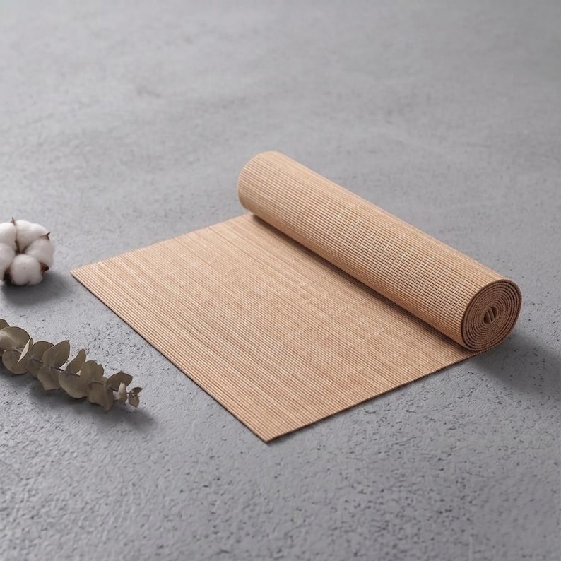 May include: A light brown, rectangular, ribbed mat partially rolled up, accompanied by a small white cotton boll and a dried eucalyptus stem, all featuring a textured surface and neutral, earthy tones for a simple, functional display.