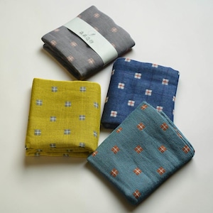 May include: Four folded square cloths in shades of gray, yellow-green, blue, and teal. Each cloth features a repeating pattern of small, cross-shaped designs in contrasting colors. One cloth is wrapped with a white band.