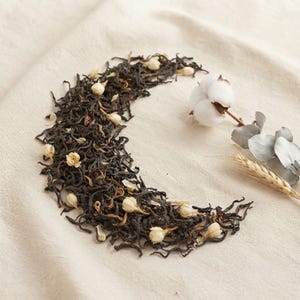 Puede incluir: Una forma de media luna formada por té de hojas sueltas de color marrón oscuro mezclado con pequeños capullos de flores de jazmín secos de color crema. Una cápsula de algodón blanca, una rama de hojas de eucalipto y una espiga de trigo dorado.