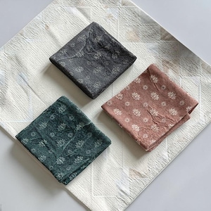 May include: Three folded square cloth napkins in shades of gray, dusty rose, and teal, each with a white floral pattern. The napkins are arranged on a textured white surface.
