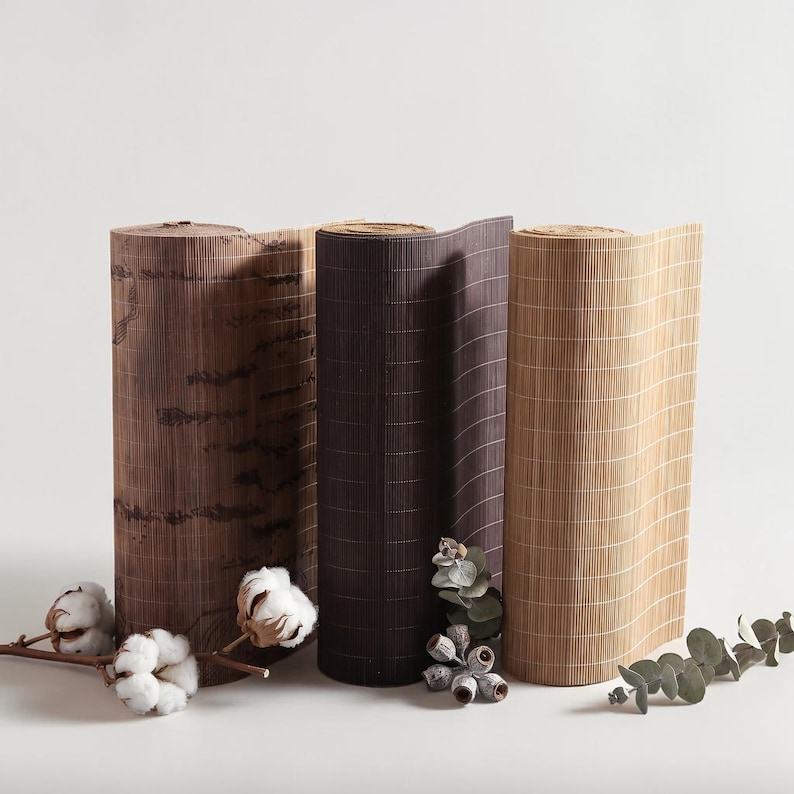 May include: Three cylindrical rolls of bamboo matting in varying shades of dark brown, medium brown, and light tan. The rolls are arranged side by side, accompanied by sprigs of white cotton bolls and green eucalyptus leaves at the base.