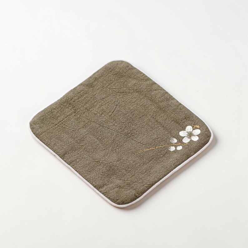 Puede incluir: Un pa&ntilde;o cuadrado verde oliva con borde blanco. Un delicado dise&ntilde;o floral en blanco y dorado adorna la esquina. El pa&ntilde;o tiene un aspecto texturizado, lo que sugiere un material suave y absorbente. Ideal para usar como pa&ntilde;o o art&iacute;culo decorativo.