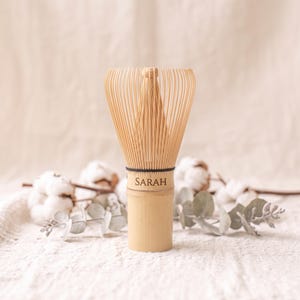 Puede incluir: Un batidor de matcha de bambú en tono beige claro con numerosas varillas finas y flexibles que irradian desde un mango central. El mango tiene grabado el nombre SARAH en una fuente serif oscura y un hilo negro fino rodea la base de las varillas.