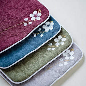 May include: Close-up of four fabric coasters in shades of burgundy, blue, green, and gray. Each coaster features a white floral design with gold accents and a white border. The coasters are stacked diagonally.
