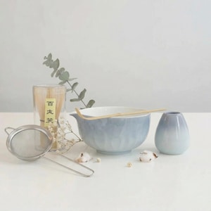 May include: A serene tea set arrangement featuring a light blue ceramic bowl with a wooden whisk, a small vase, and a glass container with noodles. A stainless steel strainer and sprigs of greenery complete the composition.